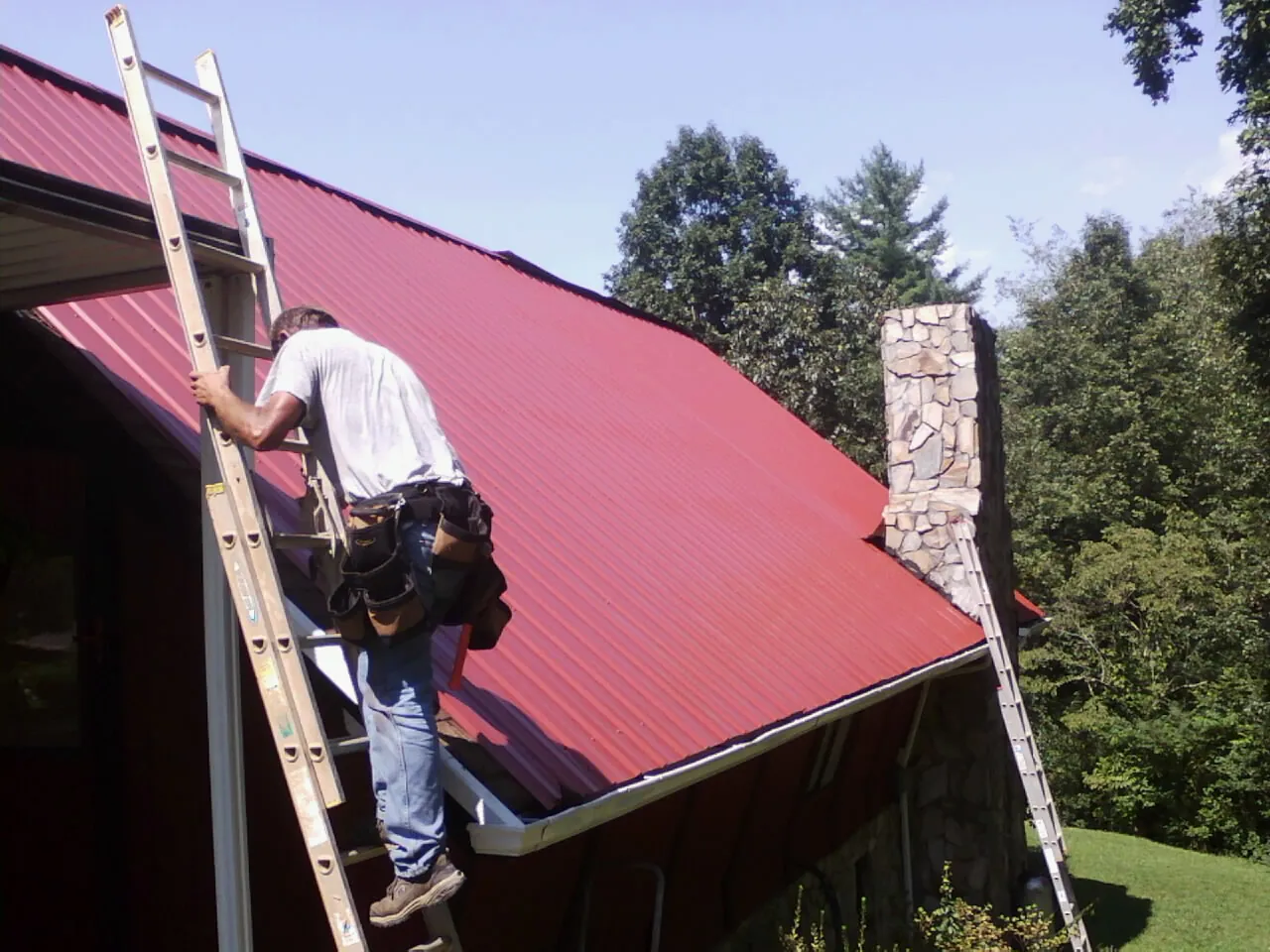 Roofer on a ladder beside a red metal roof and stone chimney — Soffit & Fascia Repair by Roofing Brothers in Menlo Park